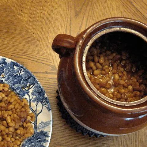 Boston baked beans in bean pot with blue plate with baked beans on the side.