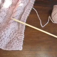 Ball of pale lavender yarn with knitting needle next to part of knitted scarf..