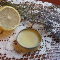 A tin of homemade solid perfume on a lace cloth with a cut lemon and sprigs of lavender beside it.