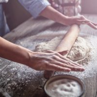 Hands using a rolling pin to roll out dough on a floured counter.