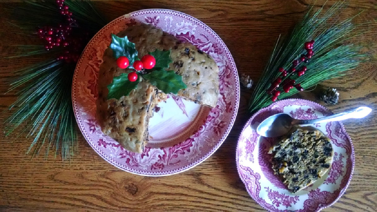 Plum pudding on a red plate with a sprig of holly in the top and a slice of plum pudding on a plate beside it and pine needles and red berries scattered around.