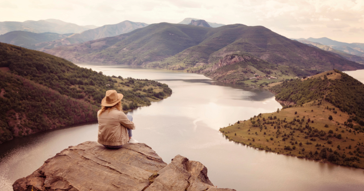 Woman sitting on top of a mountain looking out at a river and other mountains in the distance.