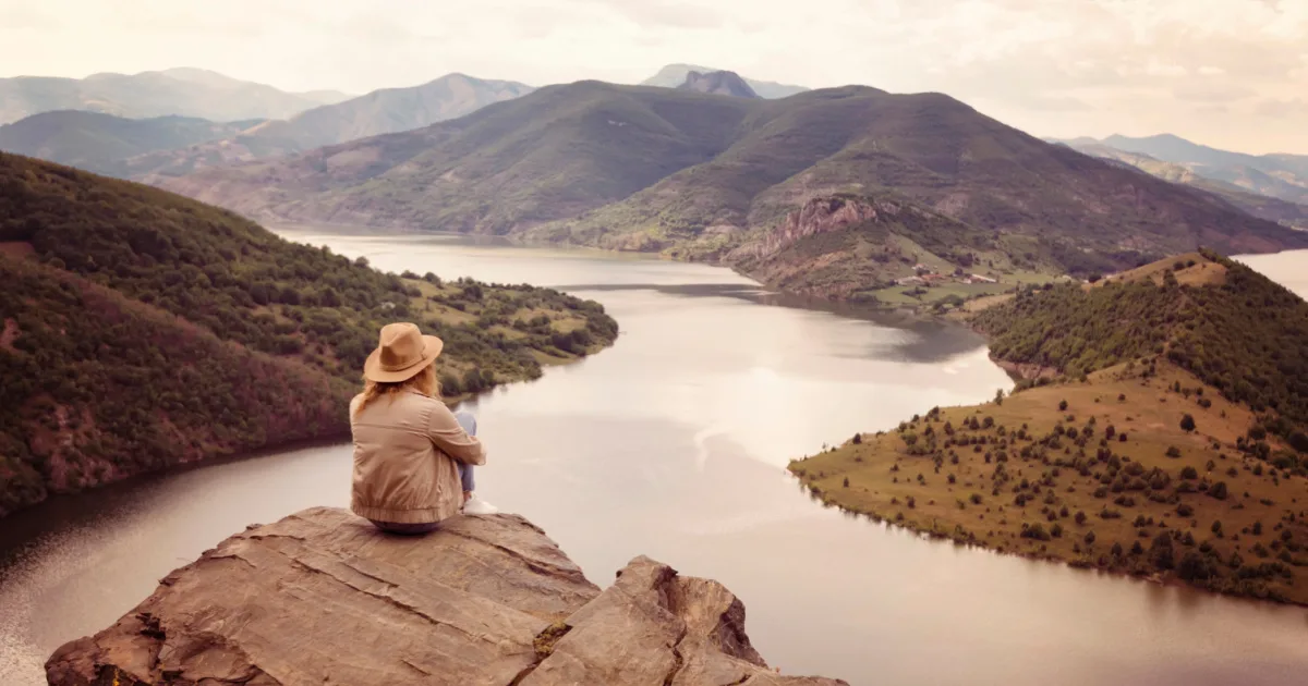 Woman sitting on top of a mountain looking out at a river and other mountains in the distance.