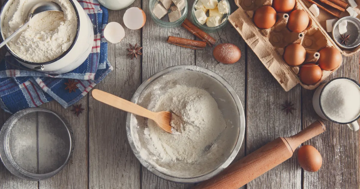 Bowl of flour on a table with eggs, a rolling pin, and other baking ingredients scattered around the table.