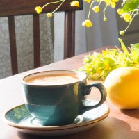 Cup of coffee in a teal blue cup with lemons and flowers on a table in the morning sunlight.