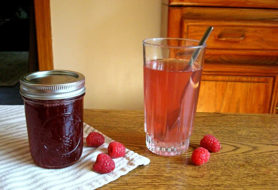 Mason jar of raspberry shrub next to a glass filled with water and shrub syrup with raspberries scattered around.