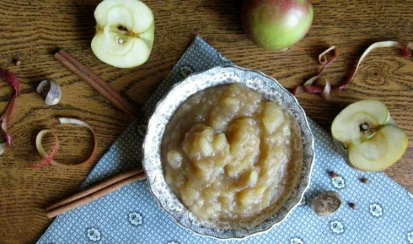 Bowl of applesauce with sliced apples and peels and cinnamon sticks scattered around.