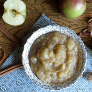 Bowl of applesauce with sliced apples and peels and cinnamon sticks scattered around.