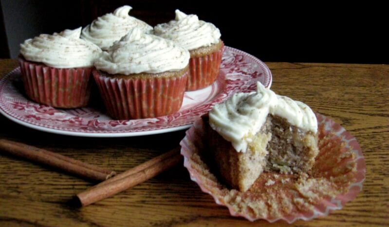 Frosted cupcakes on a red plate with a cupcake on the table beside it cut open and cinnamon sticks next to it.