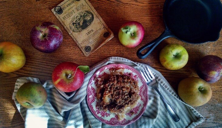 Plate of apple crisp surrounded by different colored apples and an old cookbook on a table.