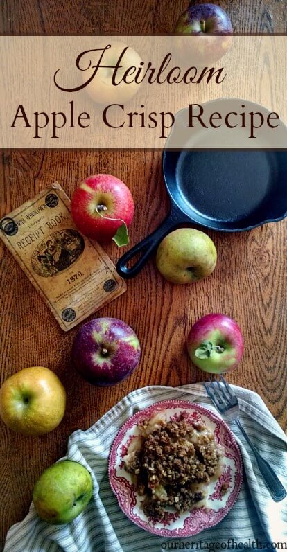 Plate of apple crisp surrounded by different colored apples and an old cookbook and cast iron skillet on a table.