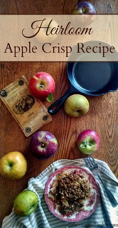 Plate of apple crisp surrounded by different colored apples and an old cookbook and cast iron skillet on a table.