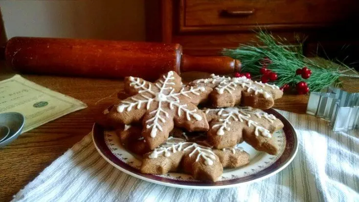Close-up images of snowflake-shaped gingerbread cookies with white icing with a rolling pin, winter pine boughs and red berries. 