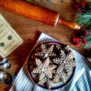 Plate of gingerbread cookies shaped like snowflakes with white icing on a table with a rolling pin, cookie cutter, measuring spoons, pine boughs, red berries, and an antique cookbook.