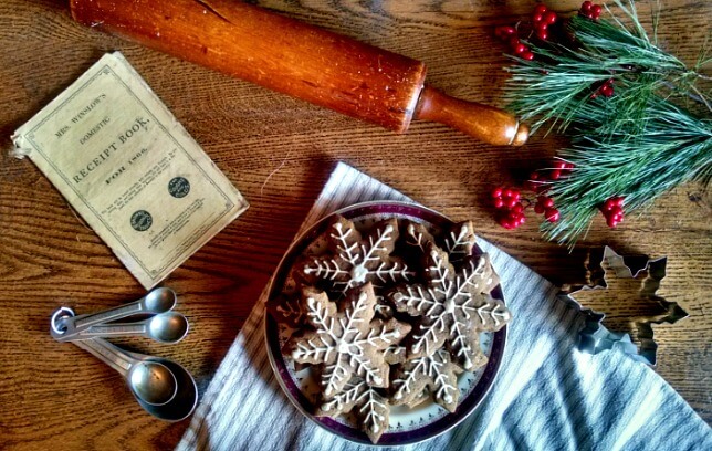 Plate of gingerbread cookies shaped like snowflakes with white icing on a table with a rolling pin, cookie cutter, measuring spoons, pine boughs, red berries, and an antique cookbook.