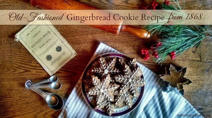 Plate of gingerbread cookies shaped like snowflakes with white icing on a table with a rolling pin, cookie cutter, measuring spoons, pine boughs, red berries, and an antique cookbook.