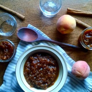 Bowl and Mason jars full of peach jam on table with peaches and spoon.