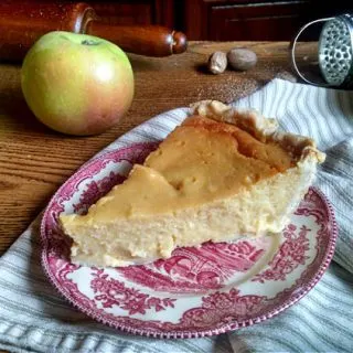 Close up of a slice of marlborough pudding on a red decorative plate with apples, a rolling pin and spice grater