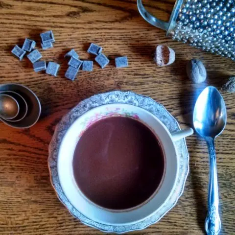 Cup of hot chocolate on a saucer with a spoon, chocolate pieces, nutmegs, a spice grater and measuring spoons on a table.