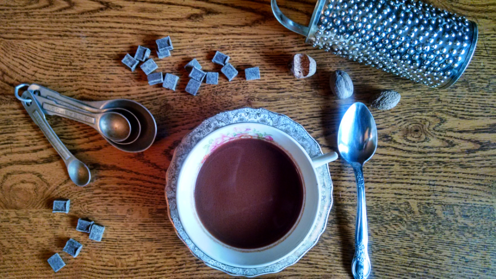 Cup of hot chocolate on a saucer with a spoon, chocolate pieces, nutmegs, a spice grater and measuring spoons on a table.