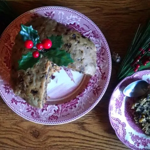 Plum pudding on a red plate with a sprig of holly in the top and a slice of plum pudding on a plate beside it and pine needles and red berries scattered around.