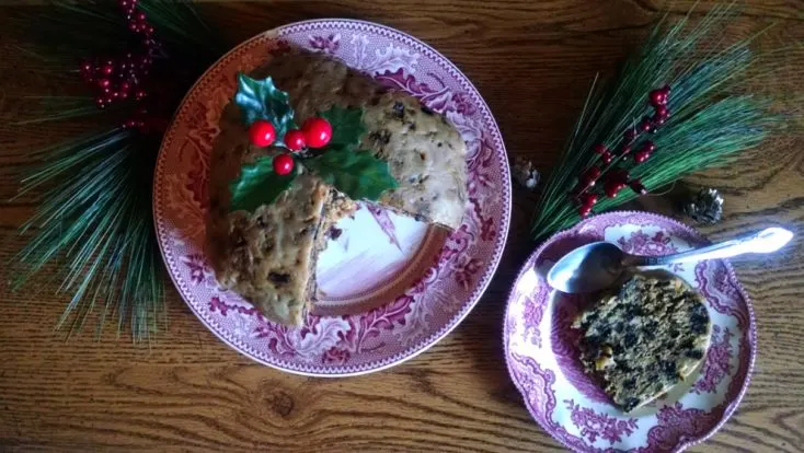 Plum pudding on a red plate with a sprig of holly in the top and a slice of plum pudding on a plate beside it and pine needles and red berries scattered around.