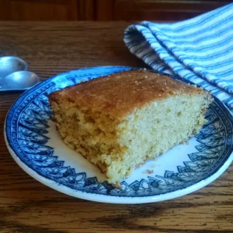Square slice of corn bread on a blue and white china plate on a table with measuring spoons and a blue and white striped towel beside it.