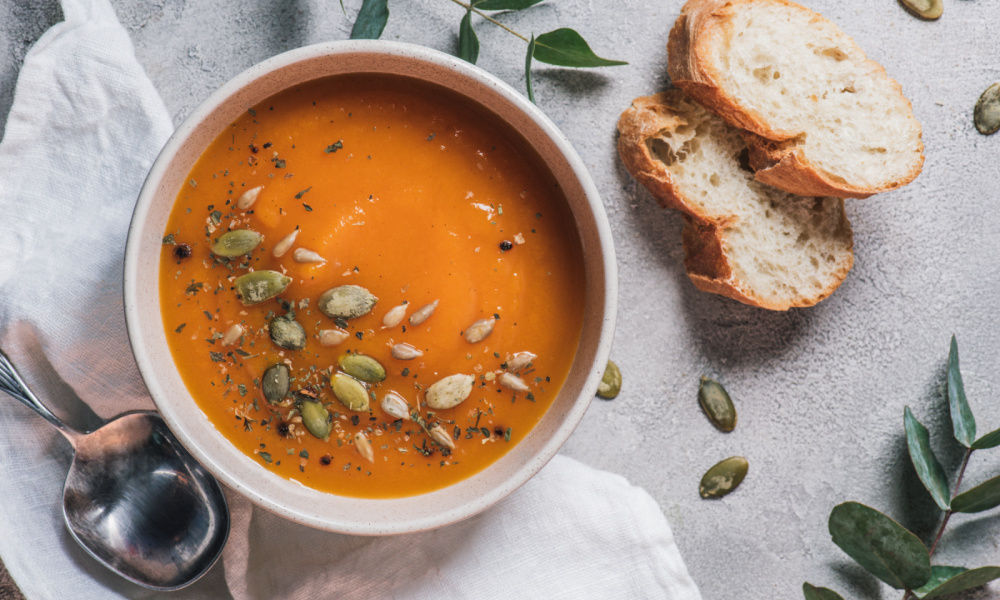 Bowl of pumpkin soup with pumpkin seeds sprinkled on top and slices of bread and a spoon beside it with leaves and pumpkin seeds scattered on the table.