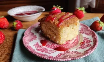 Slice of buttermilk cake with strawberry sauce on a red china plate on top of a green cloth and strawberries scattered on the table and a bowl of strawberry sauce in the background.