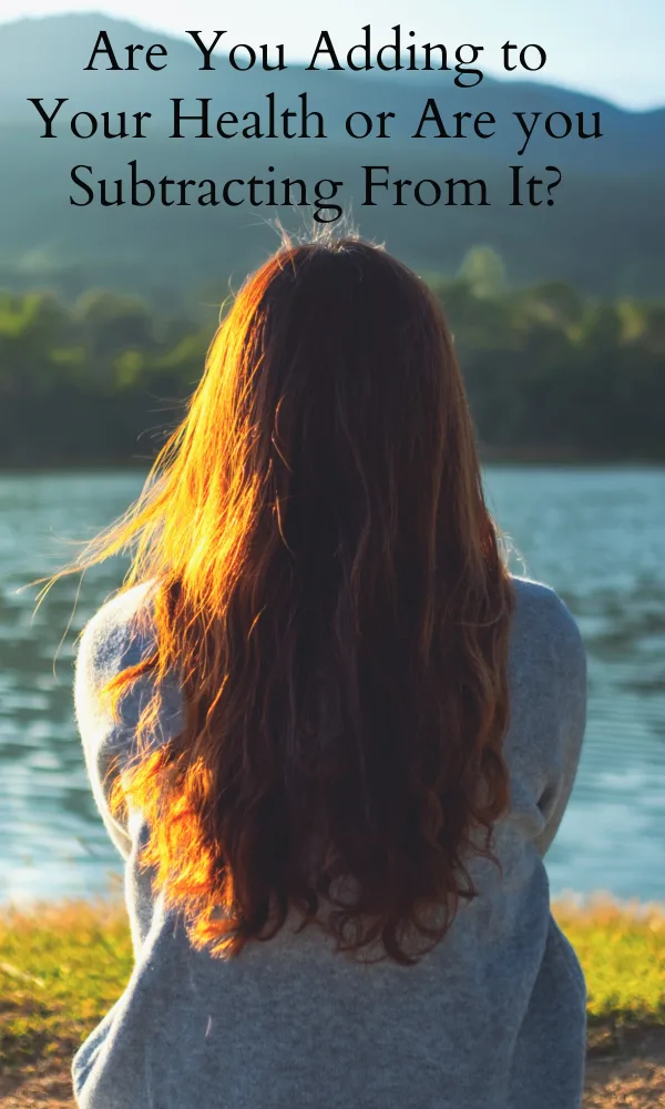 Woman sitting on the banks of a river facing away from the camera with trees and hills in the distance.