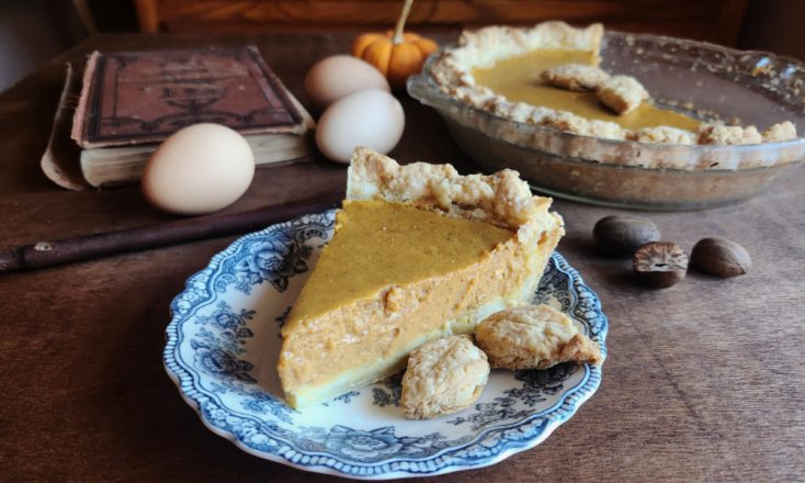 Slice of pumpkin pie on a blue china plate with more of the pie in a baking dish behind it and an antique cookbook, a small gourd, eggs, a spoon, and nutmegs on the table.