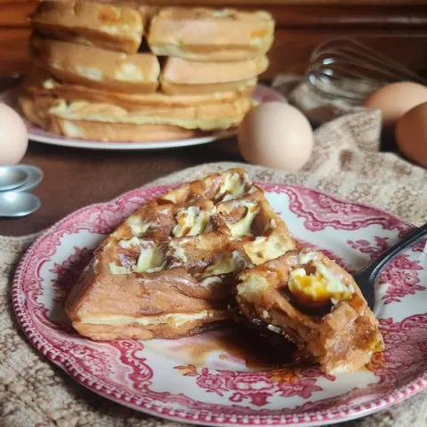 A piece of waffle with butter and maple syrup and a bit of waffle on a fork all on a red china plate with a brown striped cloth underneath and a plate piled high with waffles in the background along with eggs, a whisk, and measuring spoons.