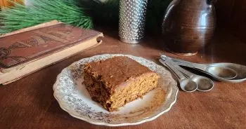 Piece of gingerbread cake on a china plate on a table with measuring spoons, an antique cookbook, winter greenery, a spice grater, and a little glazed pitcher.
