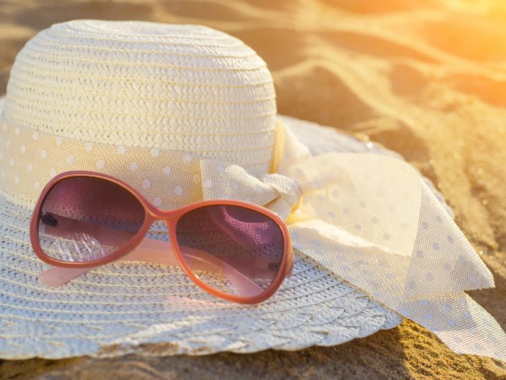 Pair of sunglasses sitting on top of a white sun hat on a beach with the glow of the sunshine.