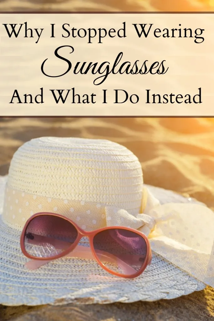 Pair of sunglasses sitting on top of a white sun hat on a beach with the glow of the sunshine with a text overlay that says "Why I Stopped Wearing Sunglasses And What I Do Instead."