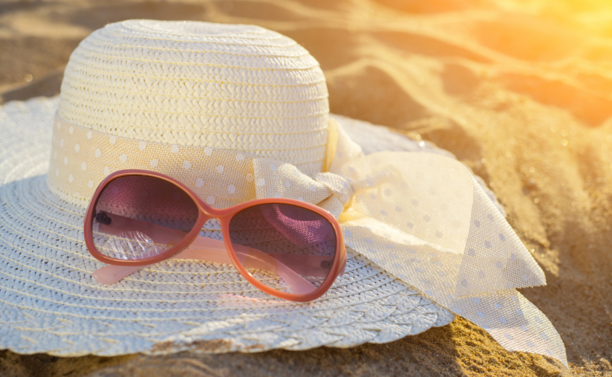 Pair of sunglasses sitting on top of a white sun hat on a beach with the glow of the sunshine.