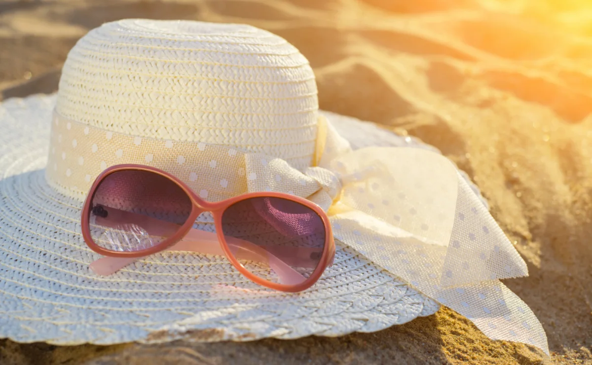 Pair of sunglasses sitting on top of a white sun hat on a beach with the glow of the sunshine.